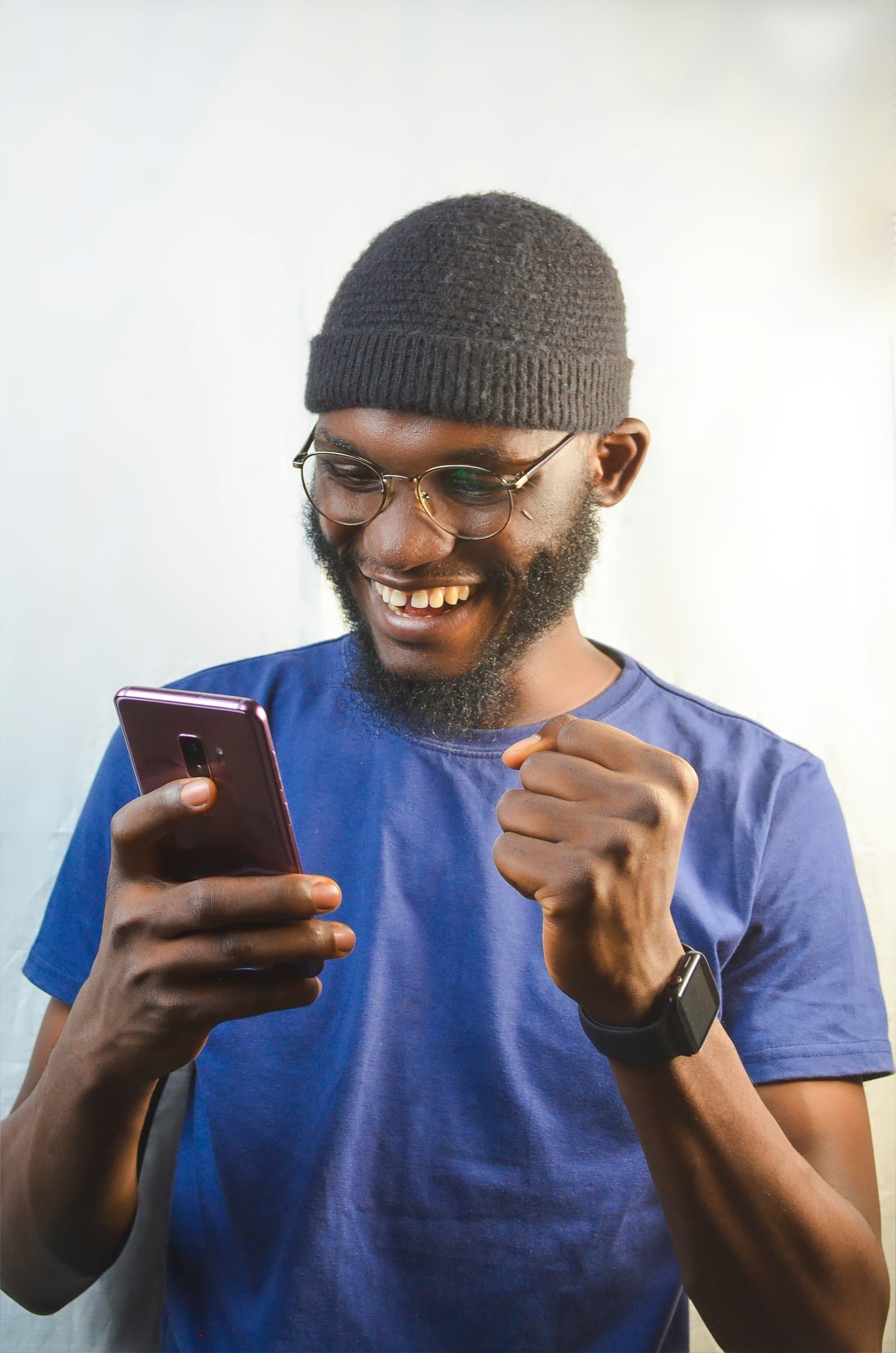 A man in glasses and a beanie grinning at his phone and pumping his fist in celebration